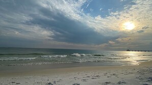 A beach with a pier extending into the water, waves gently lapping the shore, and a partly cloudy sky with the sun visible. - Seaspray Condos - 210A (Fort Walton Beach)