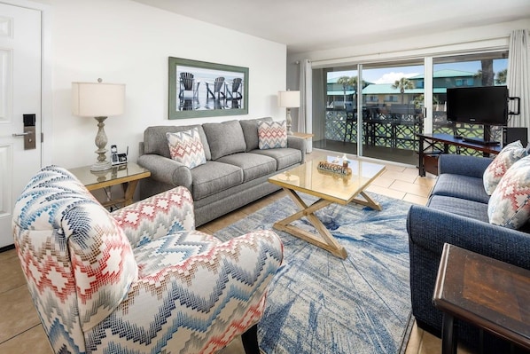 A living room with a sofa, armchair, and coffee table. There is a television on a stand and a window with a view of the outdoors. - Seaspray Condos - 210C (Fort Walton Beach)