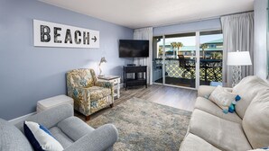 A living room with a sofa, armchair, and a chair with a 'BEACH' sign on the wall. - Seaspray Condos - 210D (Fort Walton Beach)