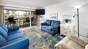 A living room with a blue sofa, a beige sofa, a patterned rug, a TV, a lamp, and a balcony with chairs.