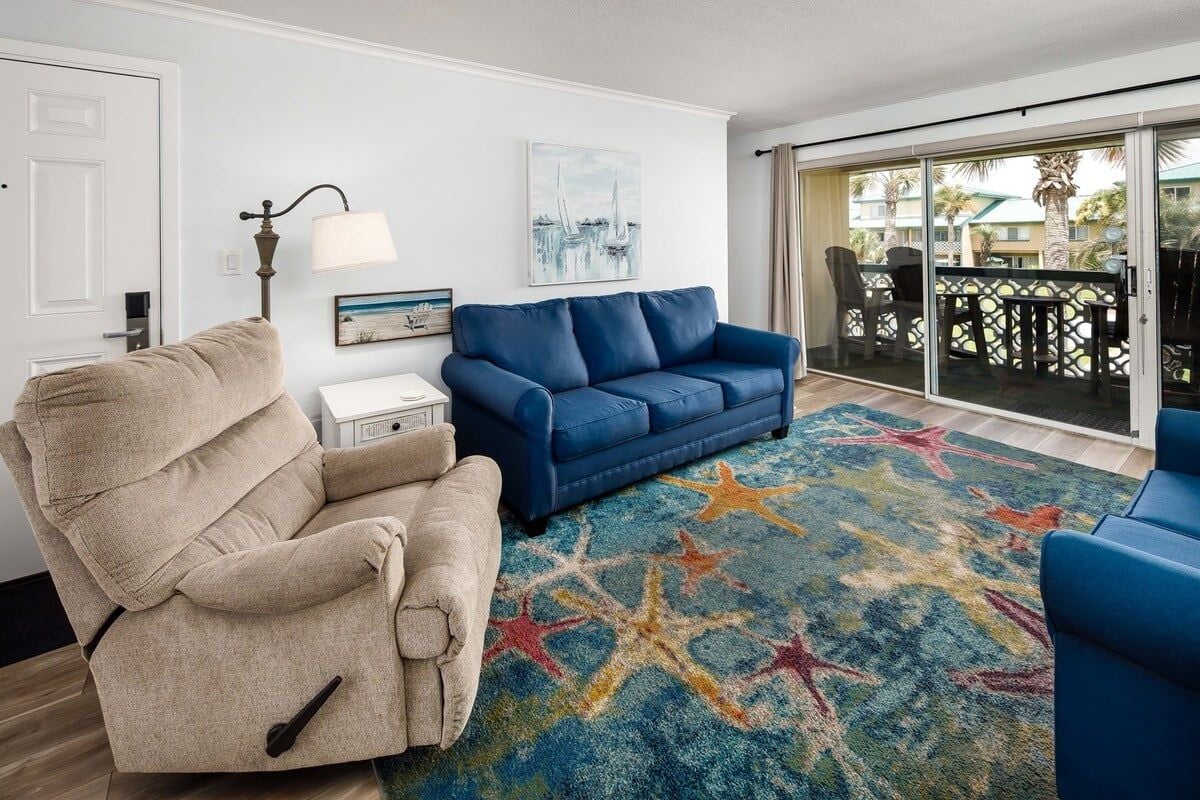 Un salon avec un fauteuil inclinable beige, un canapé bleu, un tapis à motifs étoilés et un balcon aménagé avec du mobilier d’extérieur.