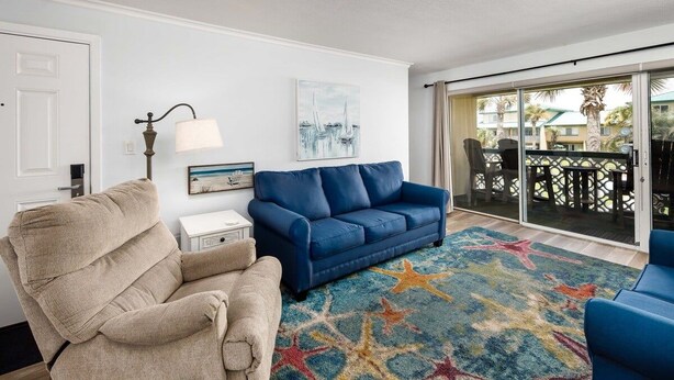 A living room with a beige recliner, a blue sofa, a star-patterned rug, and a balcony with outdoor furniture.