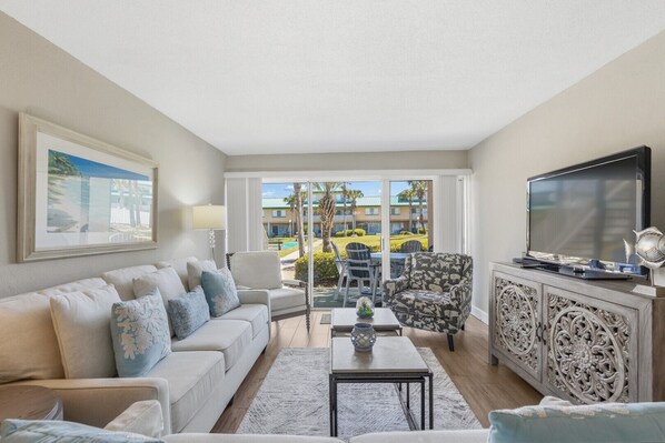 A living room with a sofa, armchair, TV, and a view of a pool and buildings.