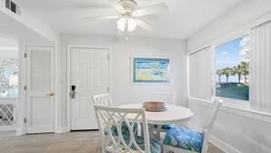 Una habitación con ventilador de techo, una mesa redonda con sillas, un cuadro en la pared y vista a palmeras a través de una ventana.