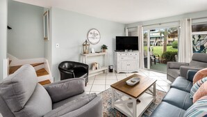 Un salon moderne avec un canapé gris, un fauteuil en cuir noir, une table basse en bois, un téléviseur à écran plat et un balcon offrant une vue sur l’extérieur.