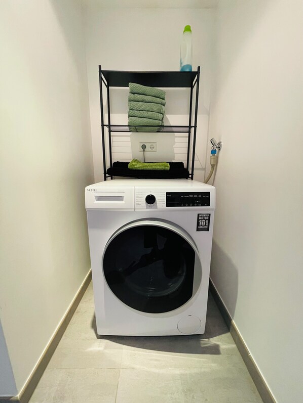 A white washing machine with a control panel, a green towel on top, and a water hose on the wall.
