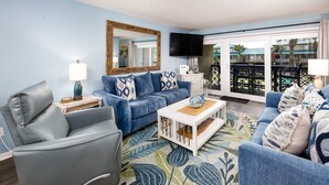 Un salon avec un canap é bleu, un fauteuil gris et une table basse blanche. Un grand miroir est accroché au-dessus du canapé et un téléviseur est fixé au mur.