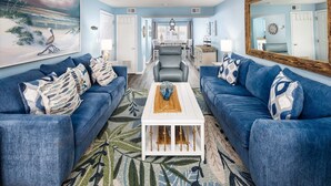 Un salon avec deux canapés bleus, une table basse blanche et un tableau représentant une scène de plage.