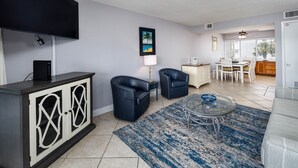 A modern living room with a flat-screen TV, a dining area, and a kitchen in the background. - Seaspray Condos - 214C (Fort Walton Beach)