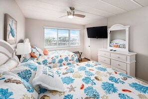 Une chambre à coucher comprenant un lit, une table de chevet, une commode, un téléviseur et une fenêtre avec des stores.
