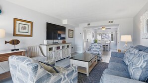 Una sala de estar moderna con un sofá, un sillón y una mesa de centro de vidrio. Hay un televisor sobre un mueble bajo y un cuadro en la pared.