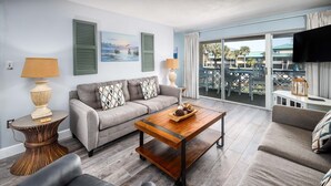 A living room with a grey sofa, a wooden coffee table, and a view of the outdoors through large windows. - Seaspray Condos - 216C (Fort Walton Beach)