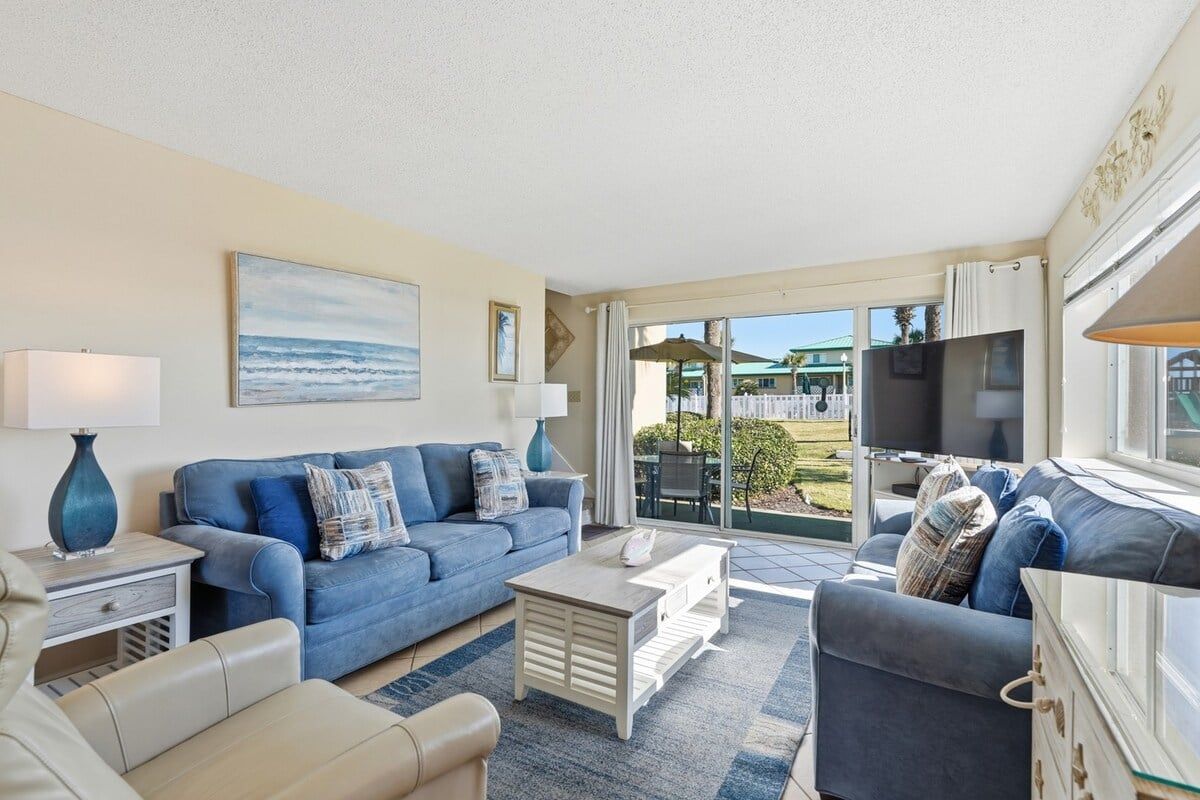 A living room with a blue sofa, a white coffee table, and a beige sofa. There is a large window with a view of the outdoors.