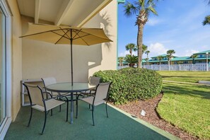 Une terrasse extérieure avec une table ronde et des chaises, un parasol beige et une vue sur un bâtiment bordé de palmiers.