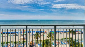 Une vue sur la plage depuis un balcon doté d’une balustrade, avec des palmiers et des parasols.