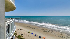 A beachfront hotel with a balcony overlooking the ocean, beach umbrellas, and a pier in the distance. - · Oceanfront King Jacuzzi Suite - Beautiful Views! (Myrtle Beach)