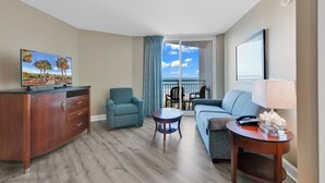 A hotel room with a wooden dresser, a sofa, a chair, a small table, a TV, and a balcony with a view of the sky.