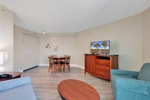 A living room with a sofa, a TV on a cabinet, a dining table with chairs, and a wall-mounted artwork.