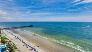 Una playa con un muelle que se adentra en el océano, una hilera larga de sombrillas de playa y un área de piscina.