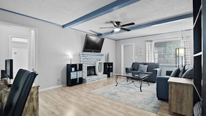 A modern living room with a fireplace, a ceiling fan, and a large window with curtains.