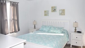Um quarto com cama, duas mesas de cabeceira, um criado-mudo, uma janela com cortinas e obras de arte nas paredes.