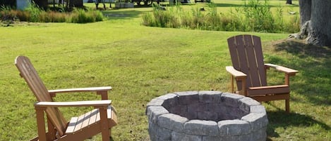 Two Adirondack chairs and a stone fire pit in a grassy outdoor area.