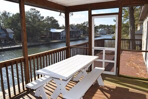 Una terraza cubierta de madera con una mesa de picnic blanca y bancos, con vista a un cuerpo de agua y casas al fondo.