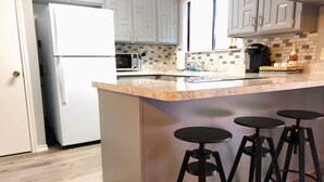 A kitchen with a white refrigerator, a microwave, a coffee maker, and a bar stool area. - •The Book Nook• Books•Arcade•Grill•Workspace! (North Richland Hills)