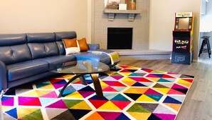 A living room with a sectional sofa, a glass coffee table, a colorful geometric rug, a fireplace, and a Pac-Man arcade machine. - •The Book Nook• Books•Arcade•Grill•Workspace! (North Richland Hills)