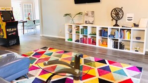 A living room with a colorful geometric rug, a Pac-Man arcade machine, a bookshelf, and a flat-screen TV mounted on the wall. - •The Book Nook• Books•Arcade•Grill•Workspace! (North Richland Hills)