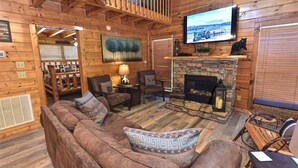 A cozy cabin interior with a stone fireplace, wooden walls, a television mounted on the wall, and a comfortable seating area with a couch and chairs. - Rustic CreekSide Memories/5min>PForge-GBurg/HotTub (Pigeon Forge)