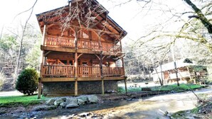 A multi-story wooden cabin with balconies, a stone foundation, and a stream in front. - Rustic CreekSide Memories/5min>PForge-GBurg/HotTub (Pigeon Forge)