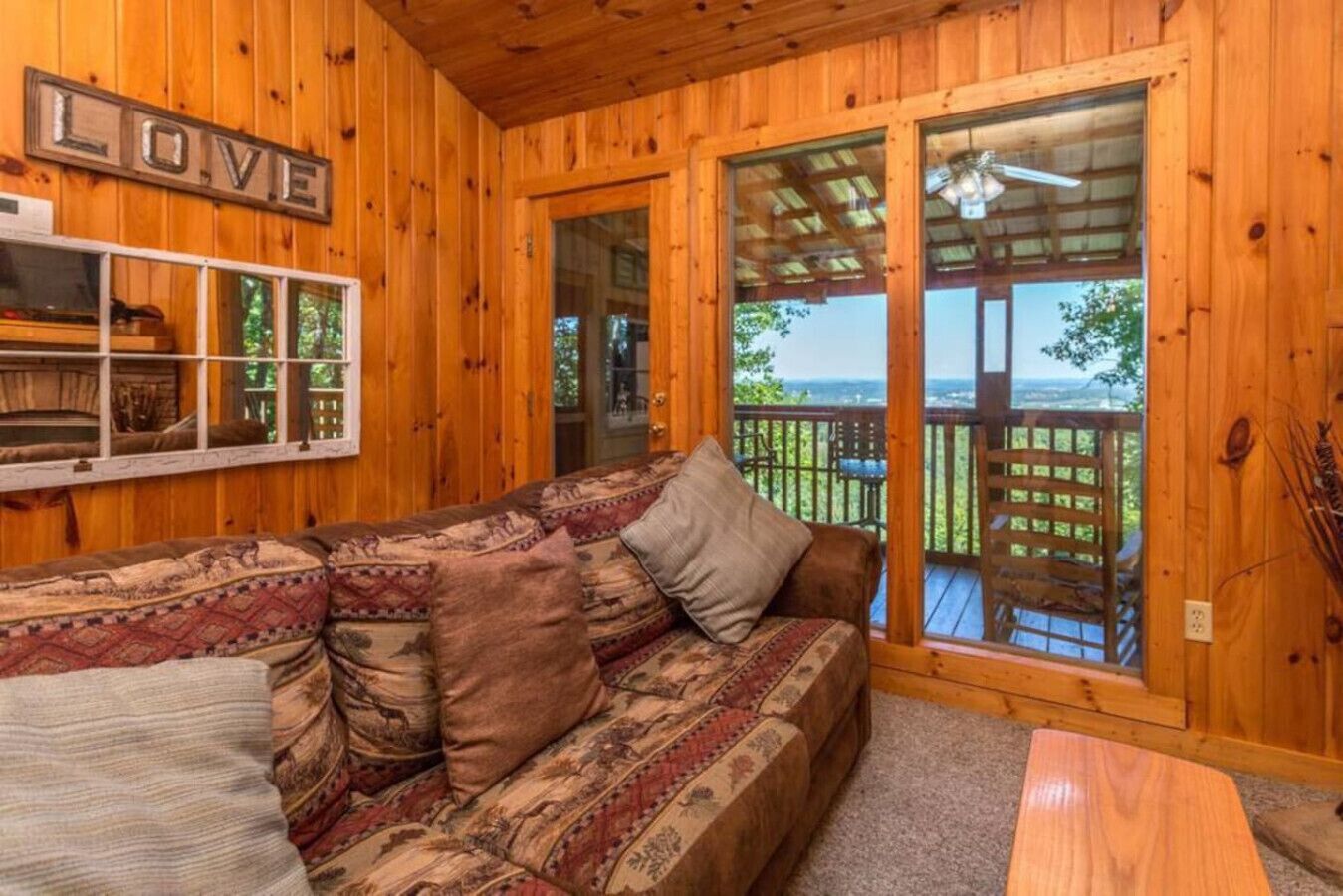 L'intérieur d'un chalet en bois, avec un canapé, un panneau décoratif portant le mot « LOVE », et une vue à travers une porte vitrée.