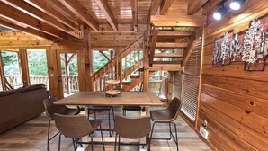 Interior de madera con una mesa central y cuatro sillas, una escalera y vistas al exterior de zonas verdes.