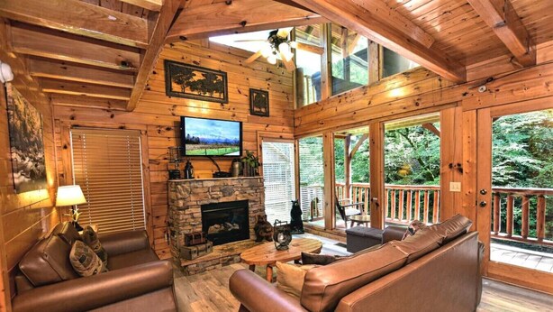Un acogedor interior de cabaña con chimenea de piedra, sofás de cuero, televisor y balcón con vistas al bosque circundante.