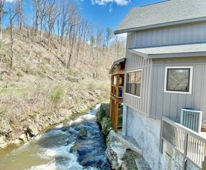 Una casa con terraza de madera que da a un arroyo, rodeada por un paisaje boscoso.