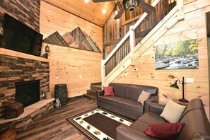 Interior acogedor de una cabaña con chimenea de piedra, paredes de madera, una escalera y un ventanal con vista al bosque.