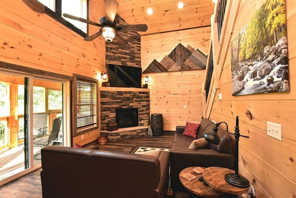 Interior acogedor de una cabaña con chimenea de piedra, paredes de madera, ventilador de techo y un cuadro de una cascada.
