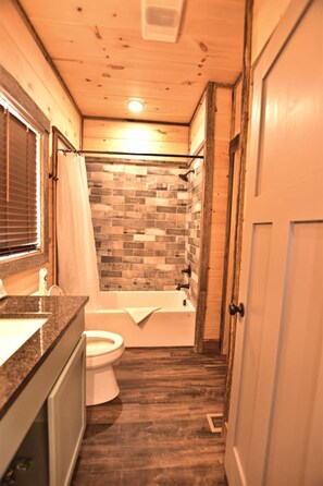 Une petite salle de bain comprenant des toilettes, un lavabo et une baignoire. Les murs sont recouverts de carreaux disposés en motif de briques, et le sol est en bois.