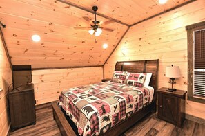 Un dormitorio acogedor con techo de madera, una cama con colcha estampada, una mesita de noche, una lámpara y una ventana con persianas.