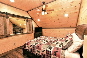 Un dormitorio acogedor con techo inclinado, una cama con colcha estampada, cabecera de madera y ventilador de techo.