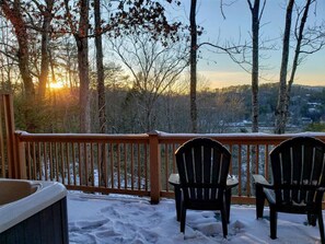 A wooden deck with two chairs and a hot tub, overlooking a snowy landscape with trees and a sunset.