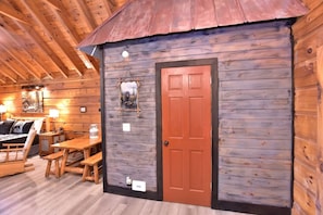 A rustic cabin interior with a wooden door, a bed, a table, and benches.