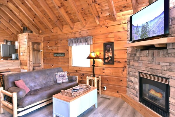 A cozy cabin interior with a wooden ceiling, a fireplace, a television, and a couch with a cushion.