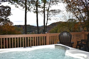 A hot tub with a wooden deck and chairs, surrounded by trees and a mountainous backdrop.