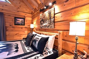 A wooden cabin room with a bed, two lamps, a framed picture, and a painting on the wall.