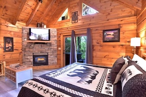 A cozy log cabin bedroom with a stone fireplace, a television mounted on a stone wall, and a large bed with patterned bedding.