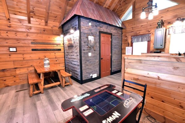 A wooden cabin interior with a small dining area, a table with a water jug, and a gaming table.