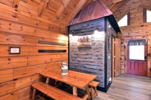 A wooden cabin interior with a rustic design, featuring a wooden table with benches, a metal pitcher, and a door with a red frame.