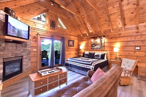 A log cabin bedroom with a fireplace, a bed with a bear-themed duvet, a wooden dresser, a sofa, and a TV on the wall.